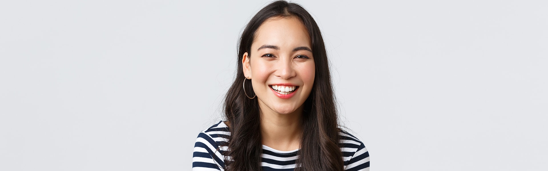 Woman with long dark hair, wearing hoop earrings and a navy-and-white striped shirt, smiling confidently in front of a plain light gray background, her radiant smile enhanced by dental crowns.