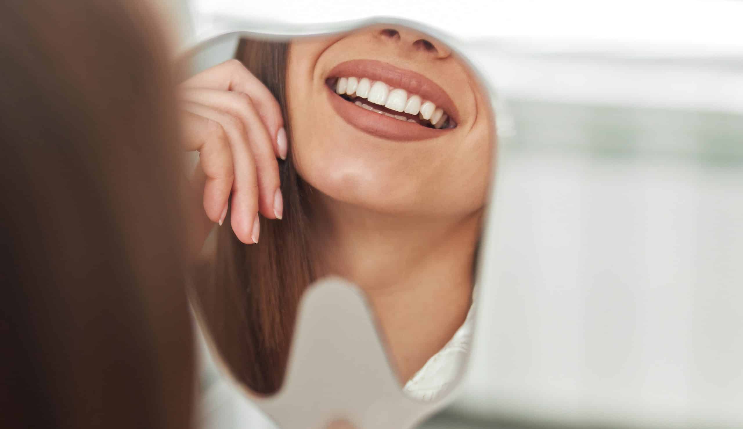 A woman’s smile is reflected in a small mirror, showing white teeth and part of her lips and chin as her hand touches her cheek—a radiant result that could only come from a trusted Wayzata Dentist.