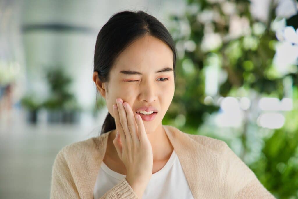 A woman touches her cheek and squints one eye, appearing to be in pain or discomfort, possibly from a toothache. She is indoors with a blurred green background, suggesting she may be in need of an emergency dentist in Wayzata.