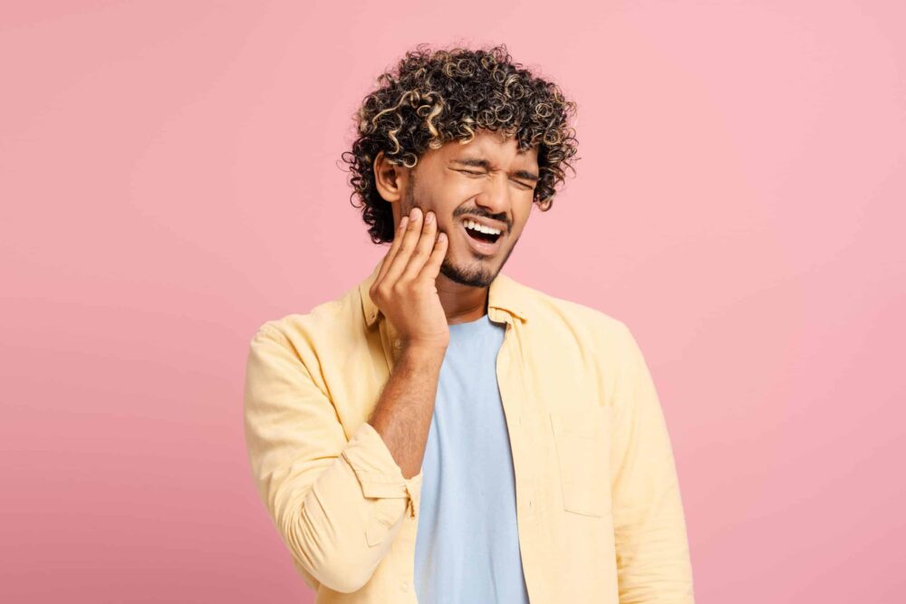A man with curly hair holds his cheek and winces in pain, suggesting a toothache that might require an emergency dentist in Wayzata. He is wearing a yellow shirt over a light blue t-shirt and stands in front of a pink background.