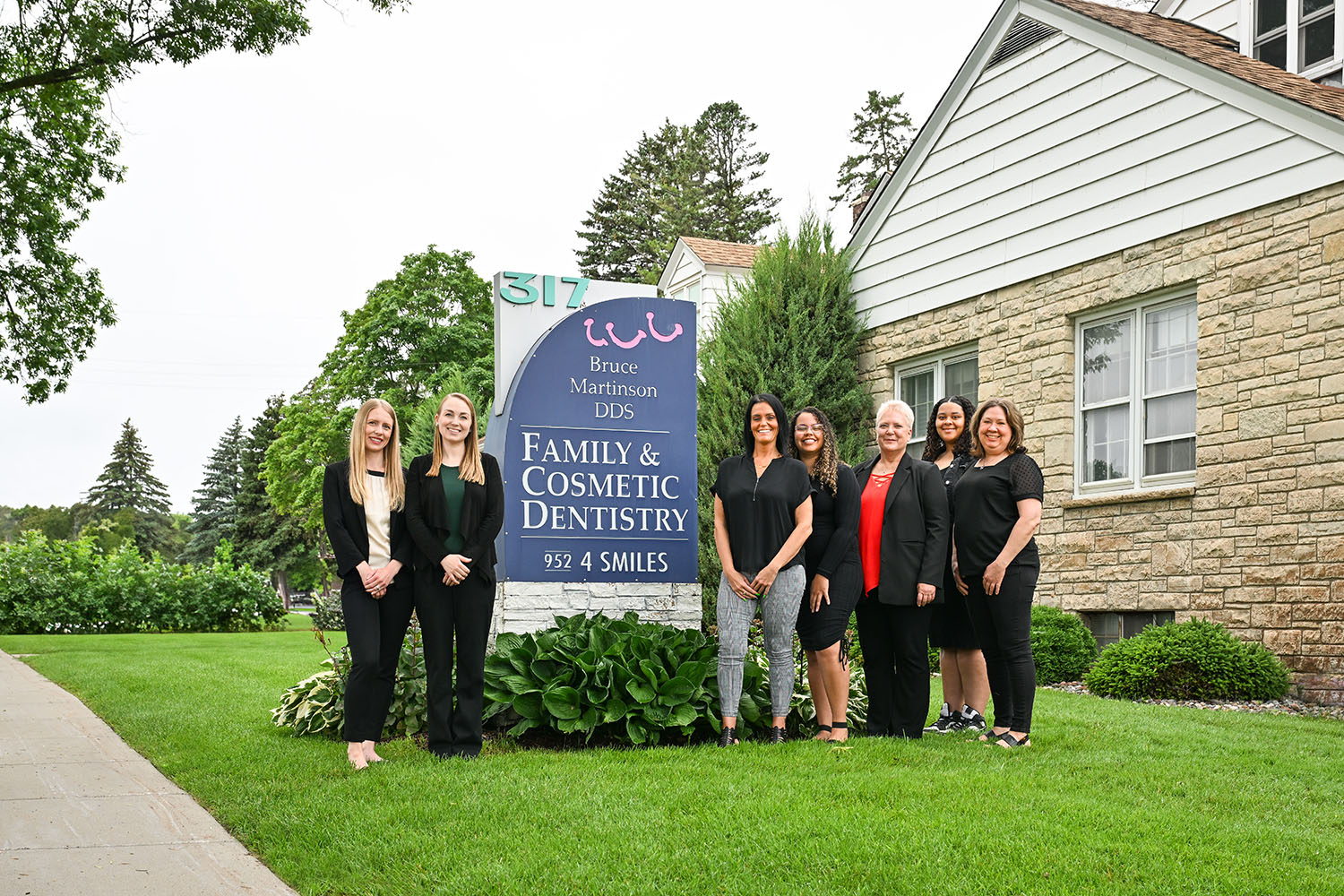 Six women stand beside a sign that reads Bruce Martinson DDS Family & Cosmetic Dentistry in front of a brick building, representing the welcoming team at Bruce Martinson DDS. Lush green grass, trees, and a sidewalk complete the inviting scene in the background.