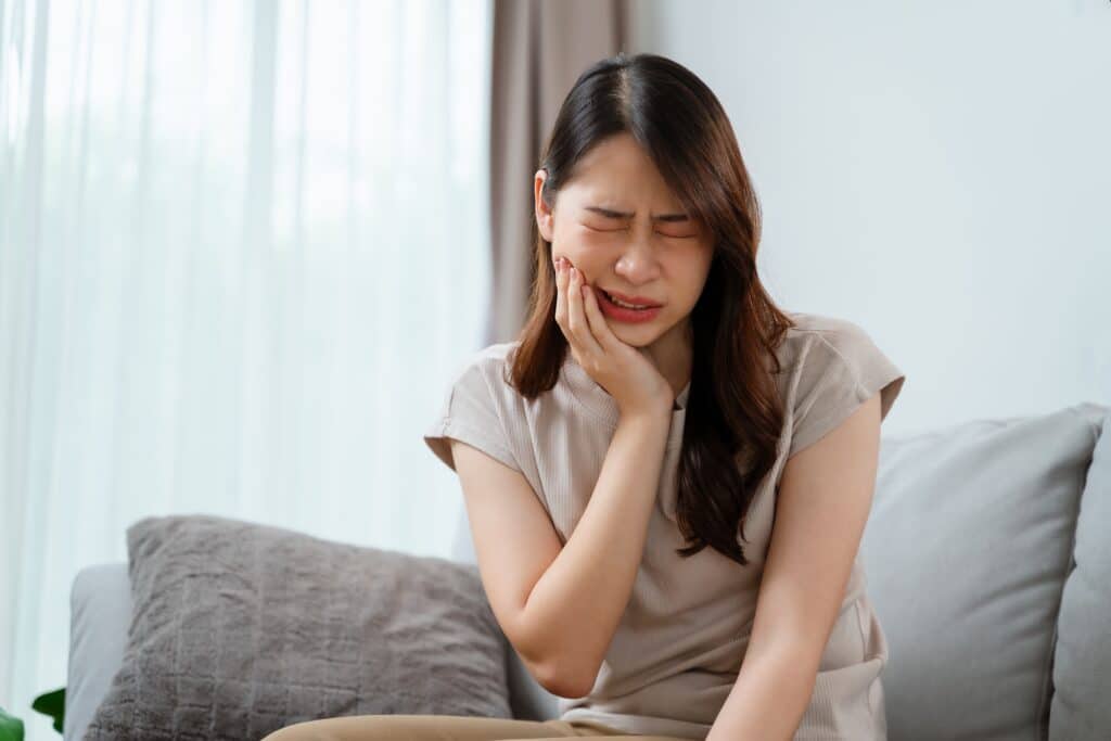 what does a cavity look like - asian woman sitting on couch with dark hair grasping her jaw in pain.