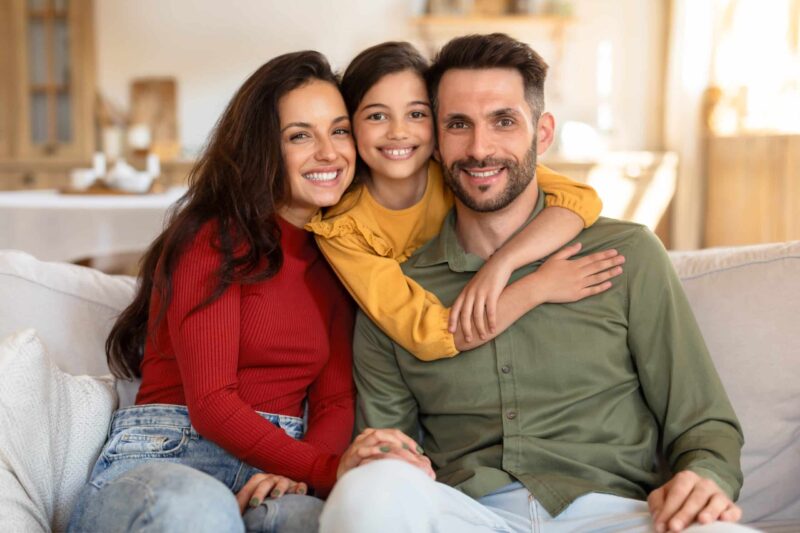 Family Dentist in Wayzata - Family of three Sitting on couch all with brown hair - mom, dad, and daughter