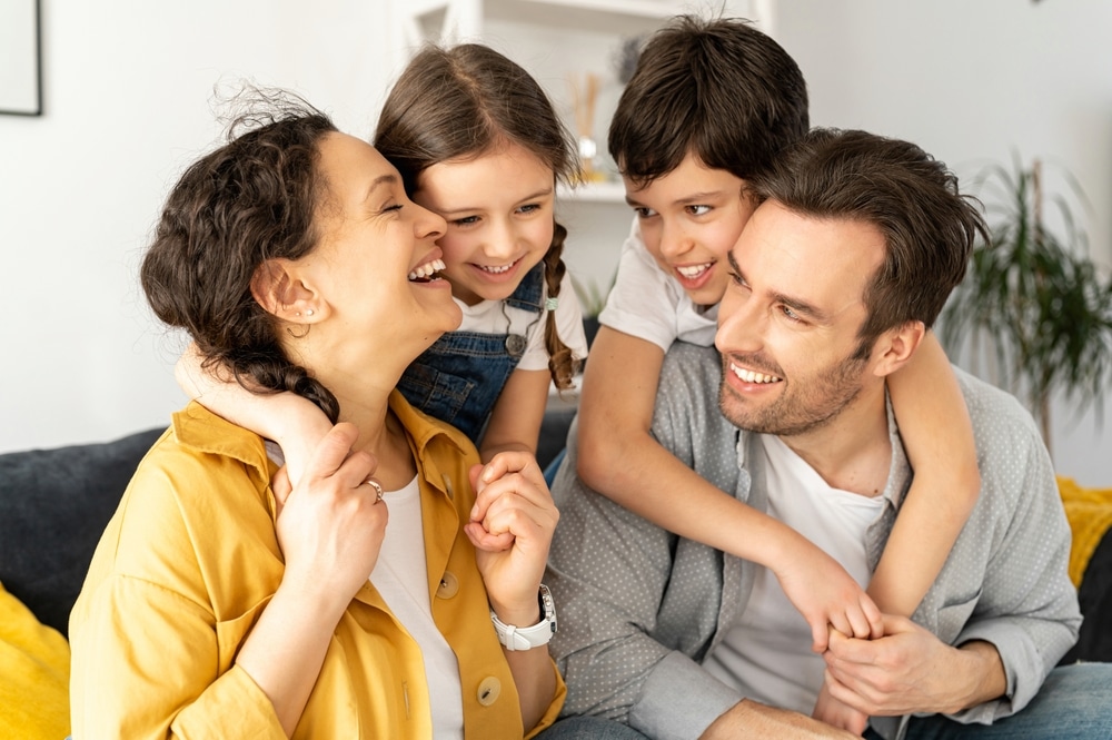 A happy family of four sits together on a couch, smiling and laughing. The two children embrace their parents, creating a warm and joyful atmosphere in a cozy living room—perhaps cherishing the confident smiles they received from their trusted dentist in Wayzata.